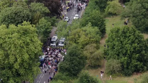 Essex Police A drone image of a large crowd of people gathered on a road, which is lined by grassed areas and trees. The people are being blocked from progressing further by police officers and their vans. More people are gathered beyond them.