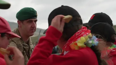 The Ten Tors Director Col Jim Bird talking to the participants who are wearing red jumpers and Hawaiian flower necklaces. 