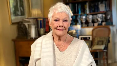 Dame Judi Dench wears a white jumper and shawl, and sits in a living room, smiling into the camera.