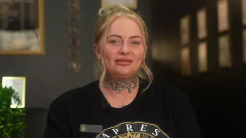 Lucy is sat in a professional piercing studio. Her blonde hair is tied up with two pieces loose, framing her face. She has a floral neck tattoo and piercings on each side of her cheeks and nose. She also has several ear piercings. She is wearing a black t-shirt with a logo on the front. An employee badge is place on the left side of her t-shirt.