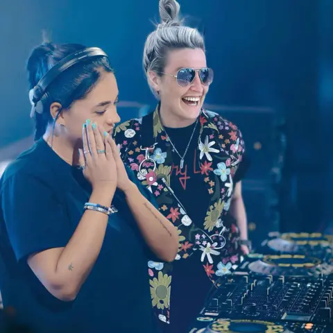 Hannah Laing A woman with black hair is wearing headphones and is standing in a black t-shirt next to DJ decks. She is next to another girl who has blonde hair and is wearing sunglasses, a floral shirt, black t-shirt and a silver necklace.
