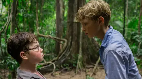 Two boys in woodland - one is very short with dark hair, red cheeks and glasses, and he is frowning, and the other is towering over him, with blond hair and a blue shirt. He looks threatening.