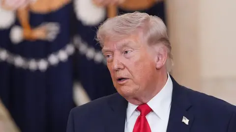 US President Donald Trump speaks while wearing a dark suit, white shirt and red tie. He stands indoors front of flag with a the US presidential seal on it.