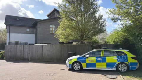 Richard Knights/BBC A police are parked side-on beside a grey garden fence. Beyond the fence is a tree and a house clad with dark wood. 