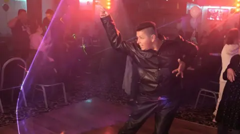 John Fairhall/BBC Taylor, who is wearing a leather jacket and trousers, is on the dance floor, pulling off a dance move.