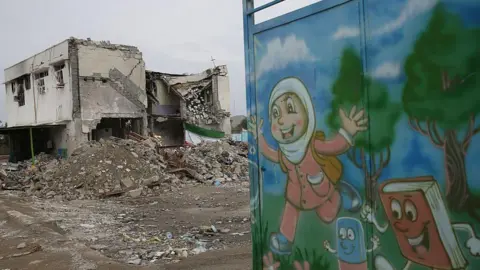 A bombed-out school surrounded by rubble, with children's illustrations painted on an adjacent gate. 