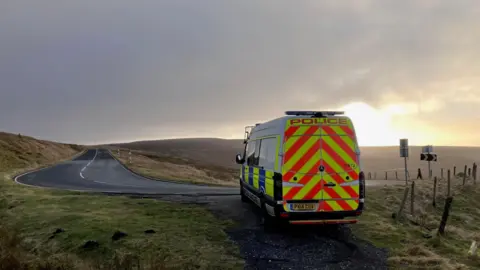Cumbria Constabulary A police van parked on a path of a bending road. The sun is setting over the hills in the distance. 