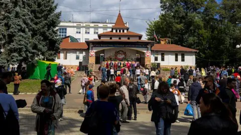 Getty Images People are standing on block paving in a city park. They are looking up the steps that lead to a white building with terracotta tiled roof and small central spire, where multiple people are standing addressing the crowd. 