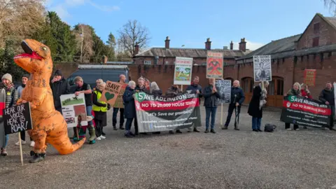 Protestors holding banners outside a council meeting. There is one person dressed as a dinosaur holding a sign saying "hear our voices, hear us roar".