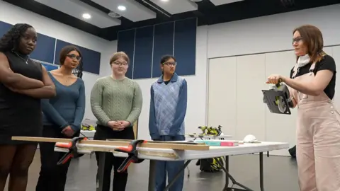 Stephen Huntley/BBC Laura Deller, on the far right of the frame, holding a circular saw and speaking to a group of four girls, who are on the other side of a table from Laura. They are standing in a line that stretches from the left of the picture to the middle. Laura is wearing pink dungarees and a black t-shirt. The girls are casually dressed.