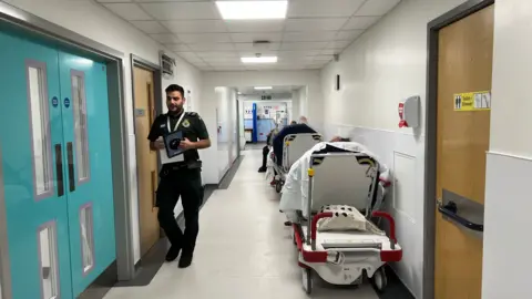 Paramedic walking down a hospital corridor with four trollies with patients waiting to be treated parked against the wall.