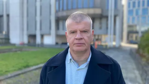 Resident John Meiklejohn standing in front of Aberdeen City Council's HQ. He is wearing a light blue shirt, and darker blue jacket. He is stood on a pavement, next to some grass.