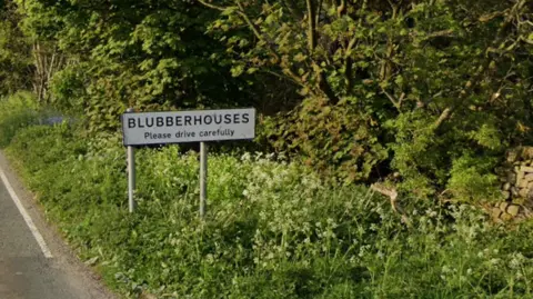 Google The picture shows a roadside sign marking the entrance to Blubberhouses, with the words “Please drive carefully” beneath the village name. The sign stands beside a narrow, rural road, bordered by tall grass and wildflowers. Behind it, there are trees and a dry‑stone wall.