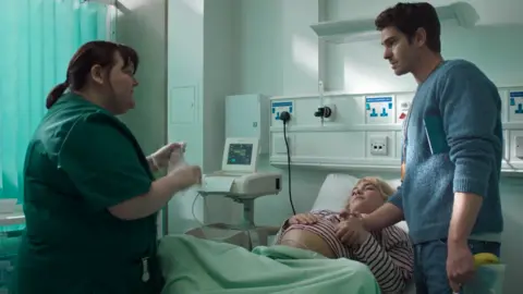 A24 Laura Guest starring as a midwife in the film A Moment In Time. She is at the end of a hospital bed - in it is Florence Pugh and Andrew Garfield stands at the top. 