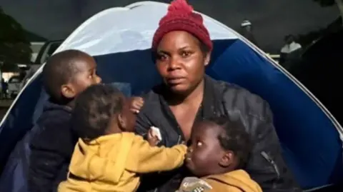 Esnat Joseph, wearing a maroon woollen bobble hat and black jacket, sitting in front of a tent with her baby triplets - two dressed in yellow hoodies and one black.