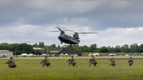 RAF Cosford A military helicopter with twin rotors hovering low above a field. Kneeling in front of it are six soldiers in camouflage and holding guns.