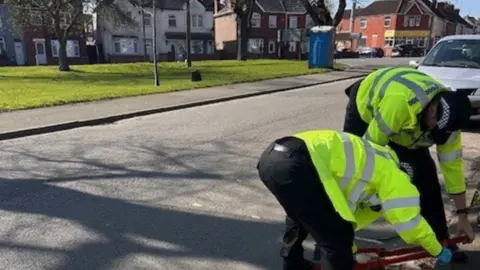 Warwickshire Police Two people in black police uniform and yellow reflective jackets bending over in a street with a grassy area on the other side of the road and a row of houses behind that