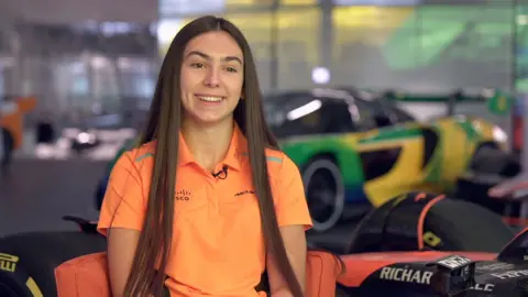 Ella Lloyd has long dark hair and is wearing a bright orange polo shirt with a McLaren logo