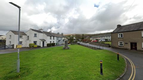 Village green in Cartmel, surrounded by a road and terraced houses. Mountains and countryside fields can be seen in the background.