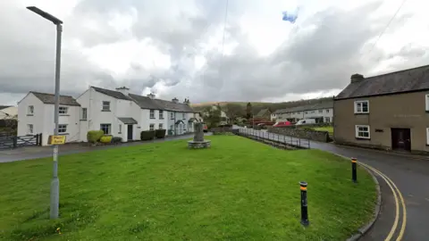 Village green in Cartmel, surrounded by a road and terraced houses. Mountains and countryside fields can be seen in the background.