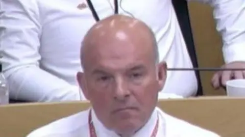 A head and shoulders image of Cllr Cutts in the council chamber. He has a shaved head and has brown eyes. He's wearing a white shirt.