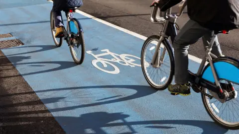 The backs of a child and an adult who are cycling on bicycles. They are riding on a blue painted part of a road which has a bike sign on it.