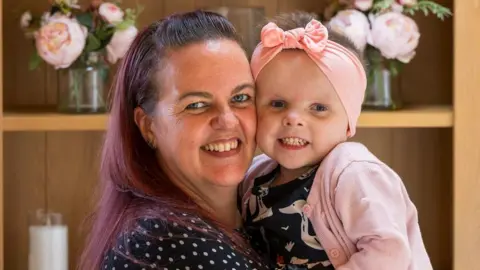 St Andrew's Hospice Sally on the left is hugging her daughter Olivia on the right - both are staring directly at the camera and smiling. Olivia is wearing a pink bow on her head and a matching pink cardigan. In the background vases of pink flowers can be seen. Sally has a black and white dress and auburn hair. Photograph was taken as part of Olivia becoming the new face of St Andrew's Hospice.