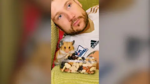 Man in his 30s pictured led on his side on a sofa, with a pet hamster infront of him leaning on a box of nuts. The man wears a white football shirt and looks away. He has a brown beard and blue eyes.