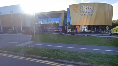 Google A view of the exterior of Camberley Leisure Centre in Surrey, from the opposite side of the road, with the sun shining over the top of the building.