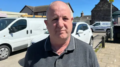 Donald's hair is cut short. He is standing outside and is wearing a chain around his neck and has a grey polo neck shirt. Behind him are a white van and white car. Another white van is parked across the street next to the brick wall of a house.
