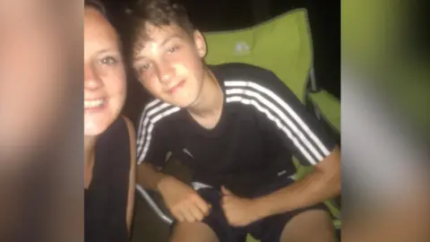 The photo shows Joshua in a black Addidas t shirt with white stripes on the shoulders. He is sitting in a green camping chair with a small smile on his face, posing for the photo next to his mum.