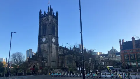 Richard Stead/BBC The photo shows a large Gothic-style cathedral with tall, pointed spires and intricate stonework. The building has a prominent clock face on its tower, and a flag is flying at the top. The cathedral is surrounded by a paved square with rows of rental bicycles neatly lined up in docking stations. In the background, modern high-rise buildings contrast with the historic architecture. The sky is clear and bright blue, suggesting a sunny day.