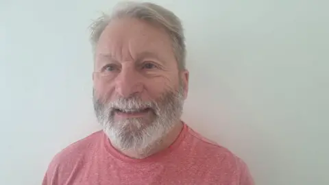A man with grey hair and a grey beard is prominent in the photo and wearing a T-shirt. He is in front of a plain, grey background.