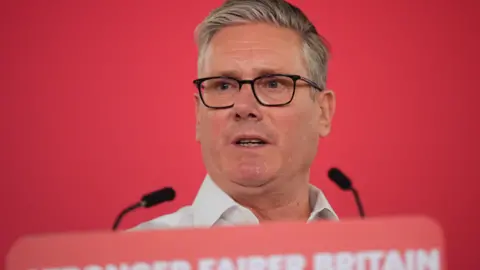 PA Media Sir Keir Starmer speaking at a podium, with Stronger Fairer Britain written along the bottom and two microphones poking out. He is wearing black rimmed glasses and an open necked shirt