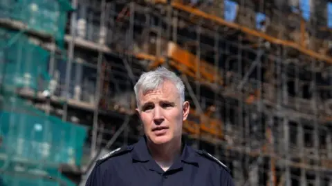 Getty Images A senior fire officer with silver hair, in uniform, stands in front of the burnt out remains of a high-rise building.