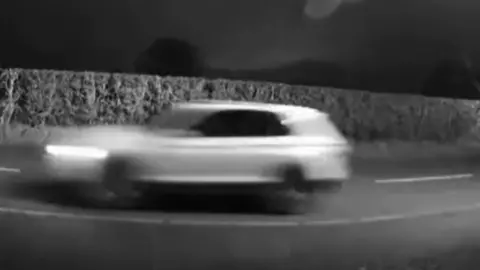 West Mercia Police An image of a white car driving alongside a hedgerow, the image is from footage captured at night and is blurred.