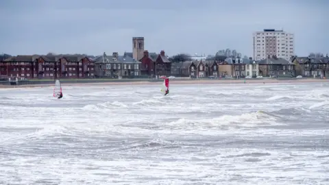 A sea coastline with two people windsurfing along the waves which are small. The weather is overcast and in the back there is houses along the seafront, and a block of flats in the corner.