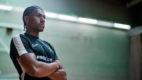 CourtesyOfZed Andre Arissol with short dark hair and beard, wearing a green T-shirt and standing with his arms folded against a green wall.