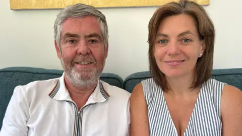 A man with short grey hair and a short grey beard with some gingery flecks wearing a white polo shirt with red and blue trim on the collar. He is sitting next to a woman with neck-length brown hair wearing a blue-and-white-striped summery dress. They are sitting on a blue sofa.