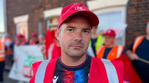 Martin Giles/BBC Gareth John, standing outside as part of a picket line. He is looking directly at the camera and there is a row of people stood behind him in the background with banners, signs and flags. He is wearing a blue and red patterned T-shirt, with a red hi-vis vest over the top and a red baseball cap on his head.