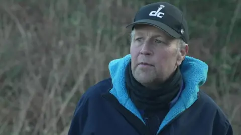 Farmer Paul Jackson who is wearing a black baseball cap, a blue coat and black turtle-neck top, is standing in a field as he is being interviewed. 
