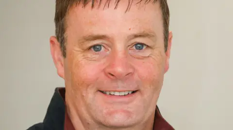 Consumer Council NI A man with short brown hair smiles and looks into the camera. He has blue eyes and is wearing navy jacket.