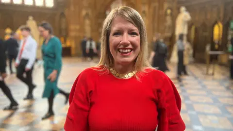 A woman with shoulder length blond hair is wearing a bright red dress and a gold necklace. She is smiling and standing in the central lobby of the House of Commons