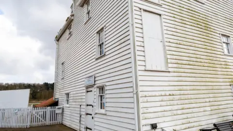 A white building that is made of white cladding stands in the middle and just off centre. It has multiple windows, and two doors.  A white fence runs from the side of the building to the left of the image. 