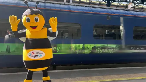 Isabella Norrison/BBC A person dressed as a bee on a railway platform, with a blue train behind them