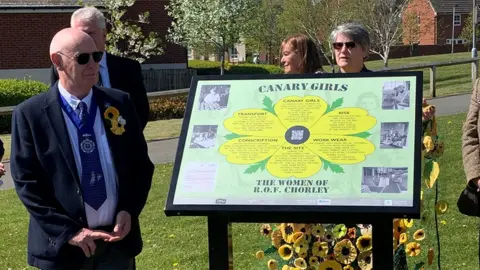 Eric Jones, who is bald and wearing sunglasses, a shirt and blue tie and dark blue suit, stands in the sunshine next to the information board which has been unveiled to show a large yellow poppy with wording on each petal and black and white phots to each side