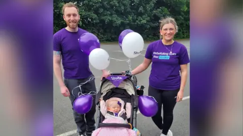 Family Anna is sitting in her pram with a pink blanket wrapped around her. She's wearing a purple bow around her head. There are purple and white balloons tied to her pram and purple bunting saying, Stroke Association on it. Anna's parents are standing next to her. Her dad is on the left and her mom is on the right. They're both wearing purple tops. One reads, rebuilding lives after strokes.