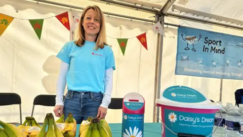 Vicky Maunder smiling at the camera has blonde hair and is wearign a turquoise t-shirt. She is standing behind a table which has bananas and water for the runners as well as a collection box.