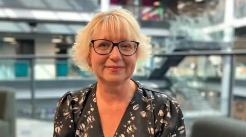 Sian has short, blonde hair in a flicky style. She's wearing black glasses and a black and white blouse. She's pictured with an office in the background.