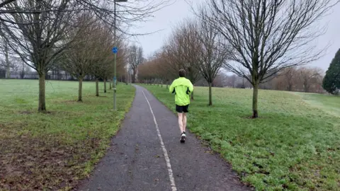 A man in a florescent yellow jacket is running down a path through a park on a misty day. Each side of the park has grass and the path is lined with trees. 